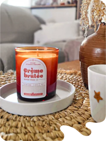 Bougie parfumée Crème Brûlée en verre ambré posée sur un plateau blanc, ambiance intérieure cosy avec vase décoratif et tasse sur une table en fibres naturelles.