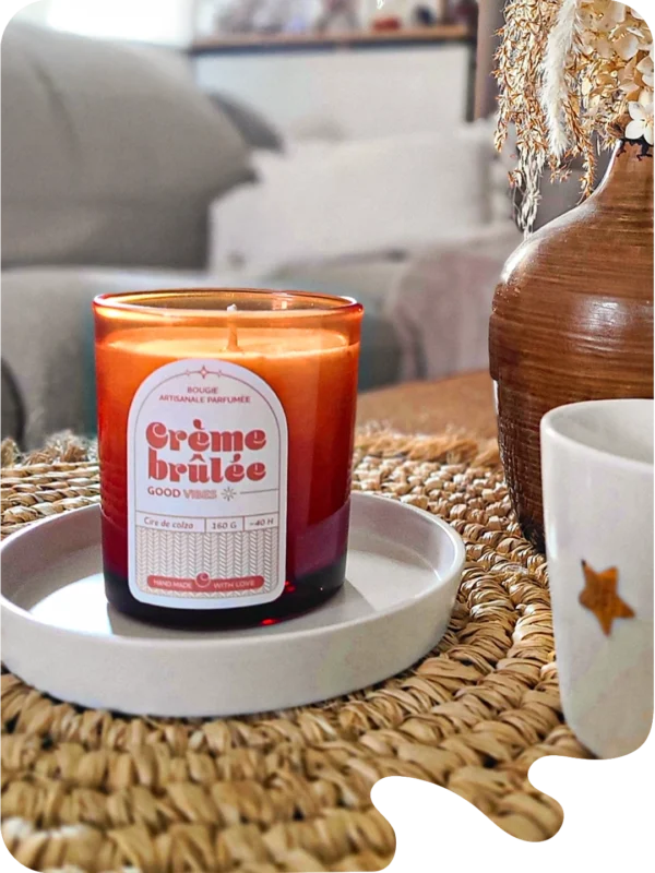 Bougie parfumée Crème Brûlée en verre ambré posée sur un plateau blanc, ambiance intérieure cosy avec vase décoratif et tasse sur une table en fibres naturelles.