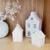 Mise en scène déco avec les bougies Home Sweet Glow en forme de maison, disposées sur une étagère en bois aux côtés d’un vase blanc et d’une lanterne céramique. Ambiance cosy et minimaliste