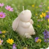 Bougie décorative en forme de lapin blanc posée dans l’herbe parmi des fleurs printanières.