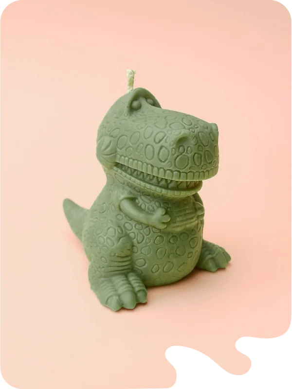 Bougie verte en forme de dinosaure T-Rex rigolo, posée sur fond violet pastel.