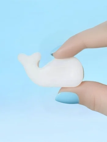 Fondant parfumé en forme de baleine tenu entre deux doigts avec vernis bleu, sur fond bleu pastel.